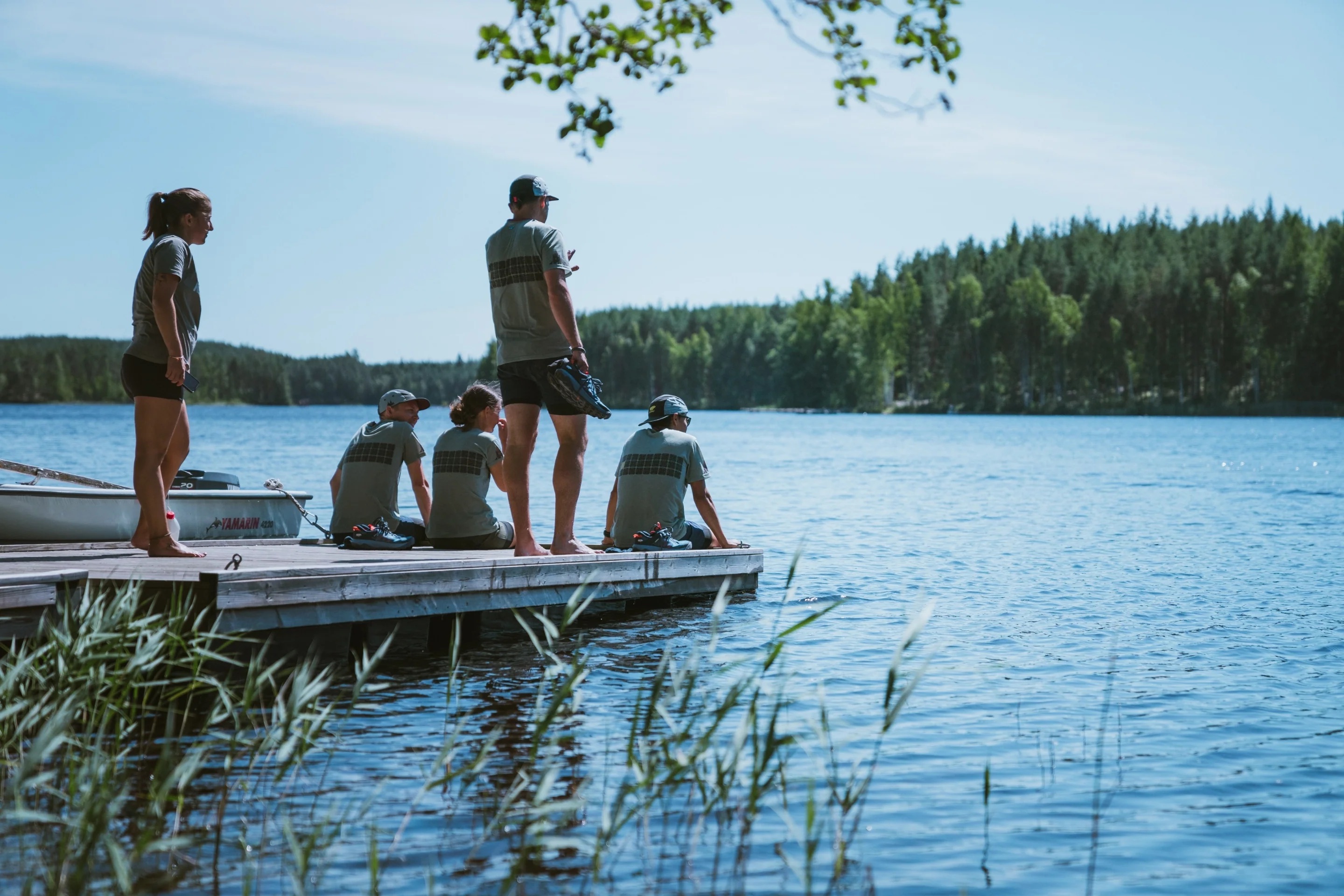 A group of people, sitting and standing, on a dock on a lake.